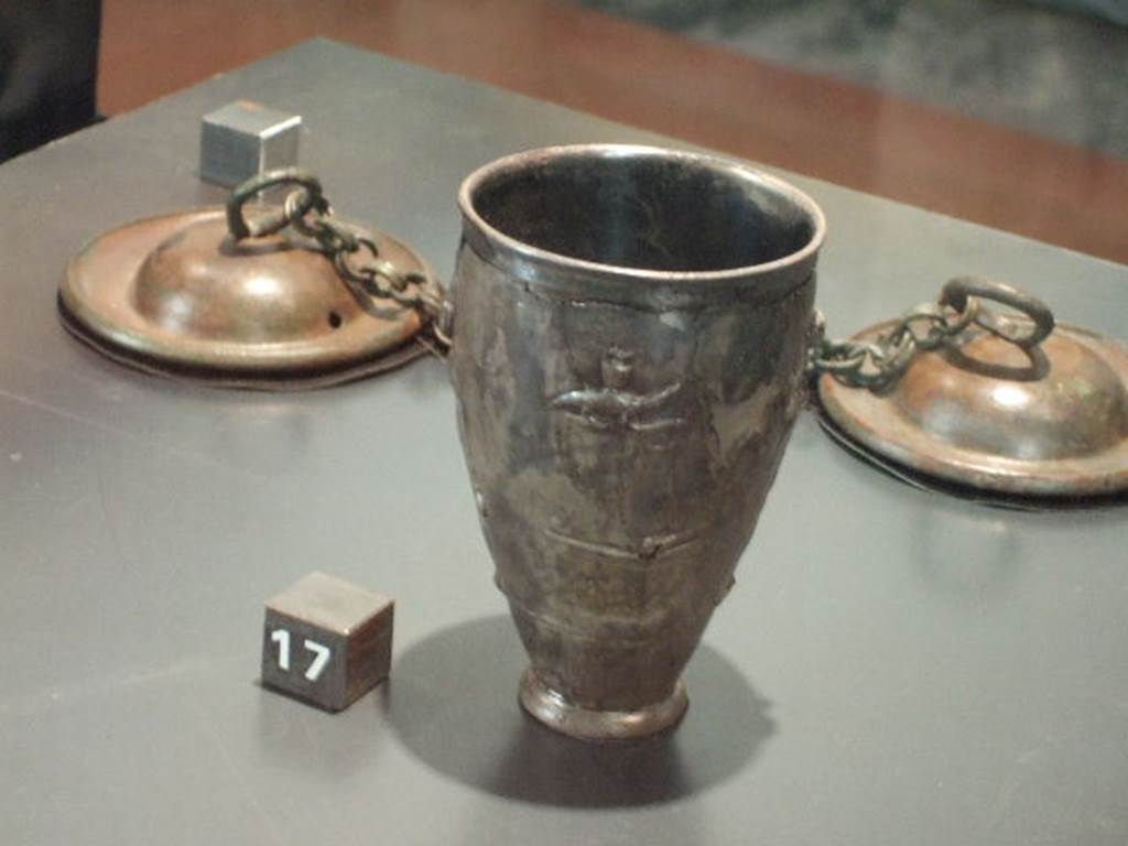 II.7.9 Pompeii. Palaestra. Silver cup with scenes of the cult of Isis. One of two found near the body of a man in the Palestra, possibly an initiate of the Isis cult. According to the Naples Archaeological Museum web site, the scene depicts the cult of Isis. A bald priest is carrying a container for holding sacred water from the Nile to the goddess Isis. Isis carries in her bent arm a statuette of the Falcon god Sokaris, depicted standing on a small base. In front of her, on an altar, there is a vase upon which there is the god Horus. An arch supported by naked Caryatids, through which it is possible to see a series of steps with palms representing the sacred area, completes the scene. These two cups represent an example of sacred, rather than household, furnishings. Now in Naples Archaeological Museum. Inventory number 6045. 