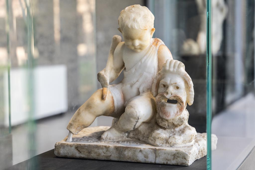 II.2.2 Pompeii. January 2023. Room “l”, (L), garden. White marble statuette of a cupid with a mask. 
Found above the marble steps in the nymphaeum at the top of the lower euripus in the garden in 1920.
On display in exhibition in Palaestra. Photo courtesy of Johannes Eber.
