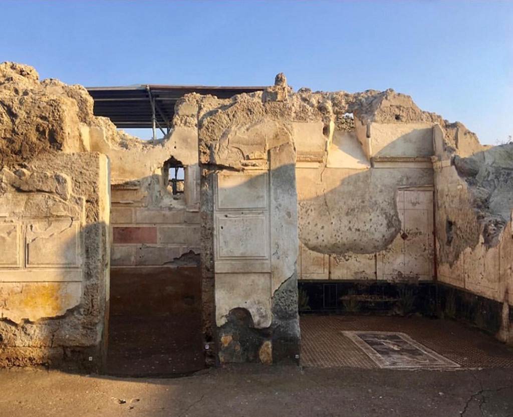 V.2.Pompeii. Casa di Orione. March 2019. 
Room A13, the south ala, on right with white walls showing Orion mosaic floor, the room was open onto the atrium.
Photograph © Parco Archeologico di Pompei.
At the time of the eruption this was a room filled with cupboards, and would have been used like the nearby room, as a storeroom.
The observation of the dynamics of entry of the pumice-stone, together with the finding of glass vases and ceramics buried inside the lapilli, at a height much higher than the floor, allow us to identify the presence of wooden furniture occupying the space (which at the beginning of the eruption had prevented the lapilli from filling the room completely). 
Also on the ground, near the two walls facing east and west there was a rich quantity of pots from table and storeroom, evidently accumulated here, due to the work in progress elsewhere. 
See Osanna M, 2019. Pompei. Il tempo ritrovato. Le Nuove Scoperte. Milano: Rizzoli, (p.101).

