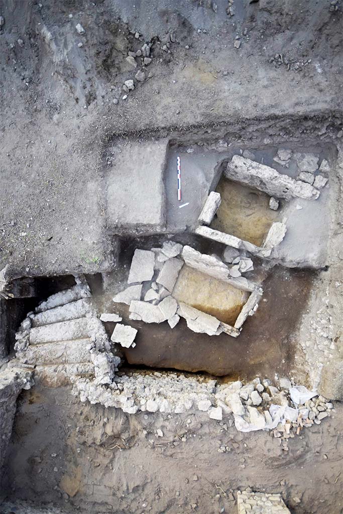 HGE31B with one side missing in 2015. HGE31A is below and the still to be uncovered HGE32A above.
HGE30 is at the bottom with the stairs of the shop at the bottom left.
Photo  B. Lemaire, EFR/CNRS.

Separated by 0.60 m from the previous one and nearly 0.50 m from the third, this grave is completely filled by reworked fill (Figs. 15 and 23). Only three Sarno limestone slabs are preserved and although the north-western one is missing, the space thus delimited is 1.44 m from north-east to south-west and nearly 0.75 m restored from north-west to south-east. The slabs measure between 0.92 and 0.99 m in height (up to 32.30 m s.l.m.) and only the one in the north-east measures only 0.62 m, but is set on wedge stones of about 10 cm. (30102). 

This burial structure was excavated in the modern period, as evidenced by the levels of fill found inside. It is only a single fill. Finally, the space observed was over-excavated by our predecessors, deeper than the installation levels, making it possible to observe the foundation of the slabs and even the wedging elements of one of them. No original floor and fill levels could be observed, nor any elements relating to the remains of the deceased or to offerings.

See CEFR 1581 or para 46 and 47 below
