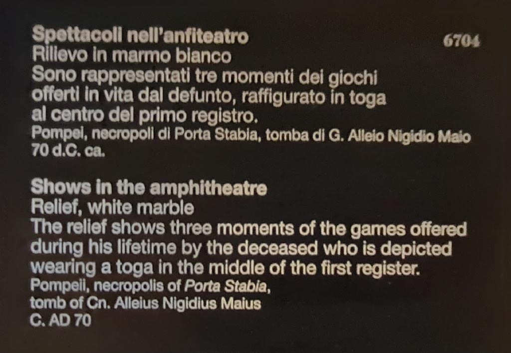 SG6 Pompeii. April 2023. Descriptive card for relief inv. 6704 in Naples Archaeological Museum. 
Shows in the amphitheatre. The white marble relief shows three moments in the games offered during his lifetime by the deceased who is depicted wearing a toga in the middle of the first row.
This card clearly identifies the tomb as being that of Cn. Alleius Nigidius Maius. 
Spettacoli nell’anfiteatro. Il rilievo in marmo bianco rappresentati tre momenti dei giochi offerti in vita dal defunto, raffigurato in toga al centro del primo registro.
Questa carta identifica chiaramente la tomba come quella di G. Alleio Nigidio Maio.
Photo courtesy of Giuseppe Ciaramella.

